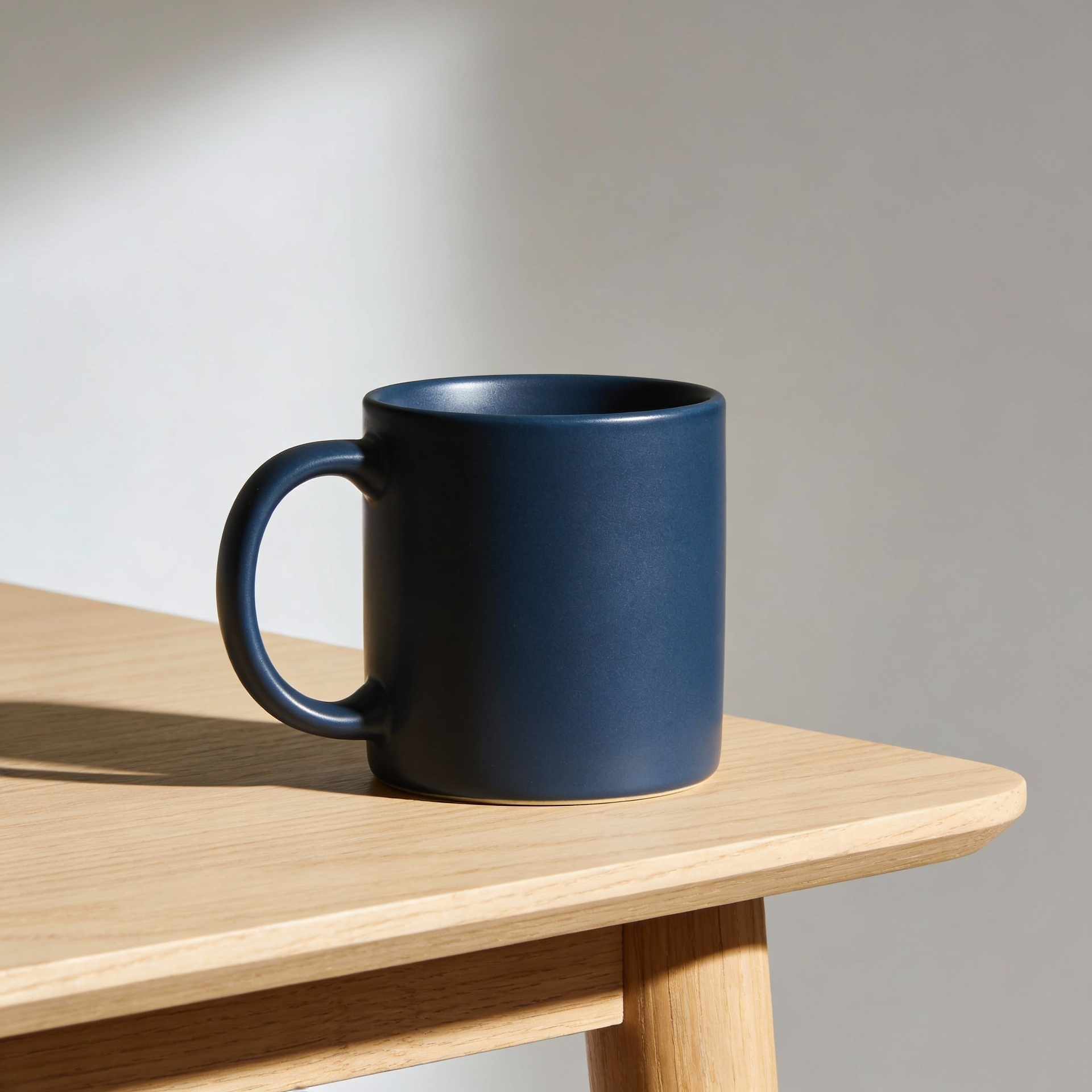 A ceramic coffee mug with a matte navy glaze, sitting on a light oak table, minimal studio setup, centered composition, 3/4 angle, softbox key light from left, gentle shadow falloff, 50mm look, photoreal product photo, clean background, must keep: navy glaze, no logo, no text.